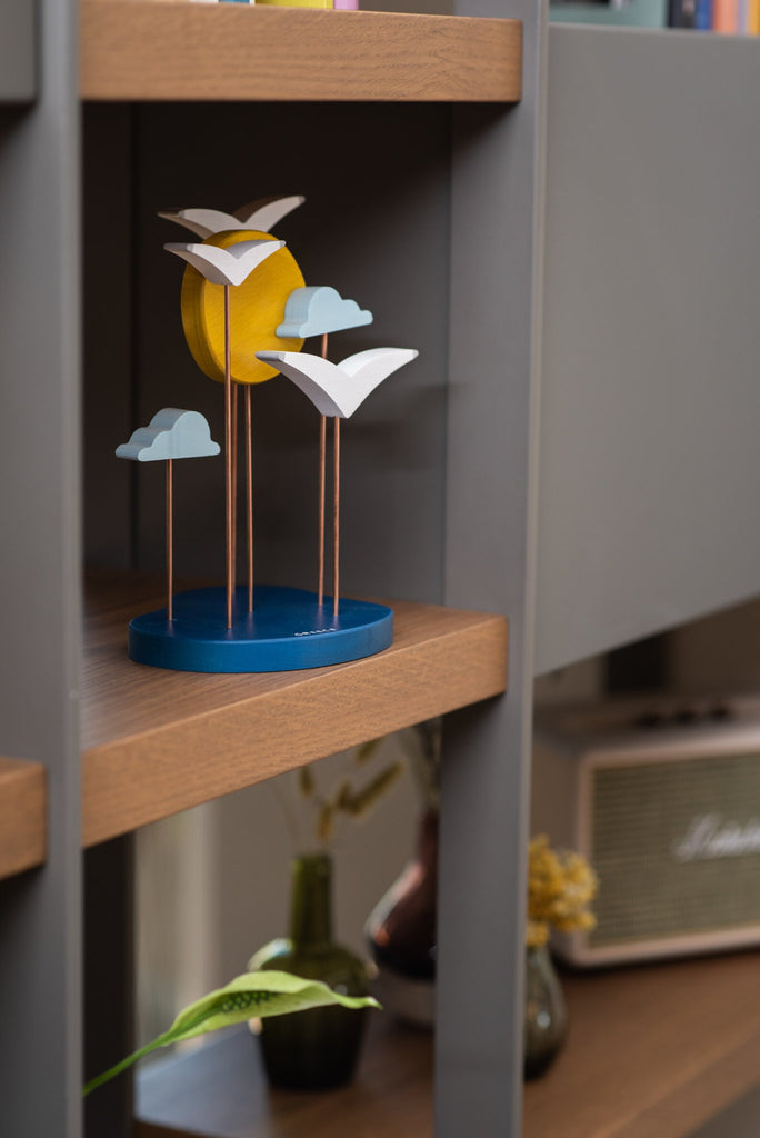A modern FSC certified European Beech Wood shelf displaying a quirky Sunset Decoration featuring a stylized sun and clouds made of metal, placed alongside books and a plant, with a speaker visible in the background