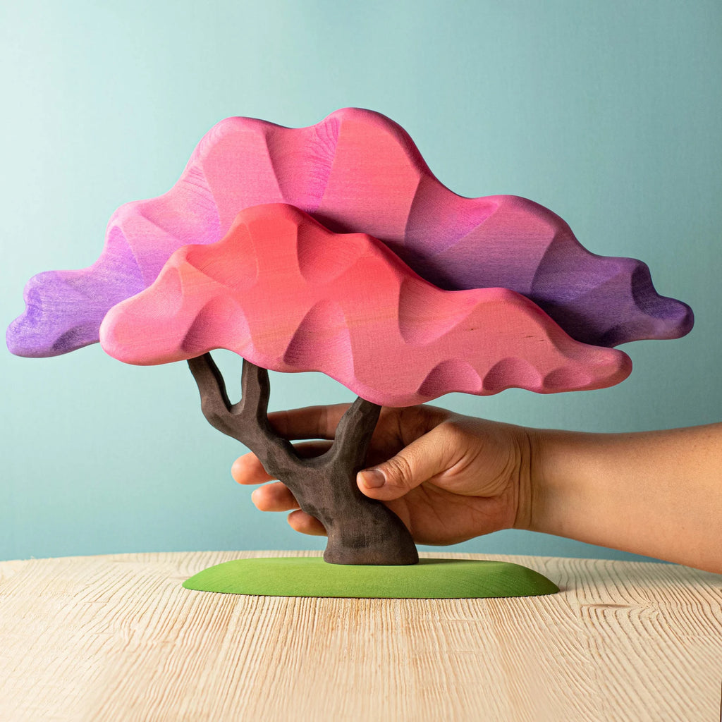 A hand holding a Handmade Japanese Maple Tree sculpture with pink and purple non-toxic paint leaves on a beige table against a light blue background.