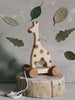 A Handmade Wooden Giraffe Pull Toy with brown spots and four wheels stands on a tree stump. The kids' toy has a white pull string attached. In the background, there is a gray wall decorated with pressed green leaves of various shapes and sizes.