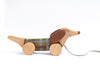 The Handmade Pull Along Dog Toy is shaped like a dachshund with large round wheels, a plaid body midsection painted with natural milk paint, and a string attached to its nose, set against a plain.