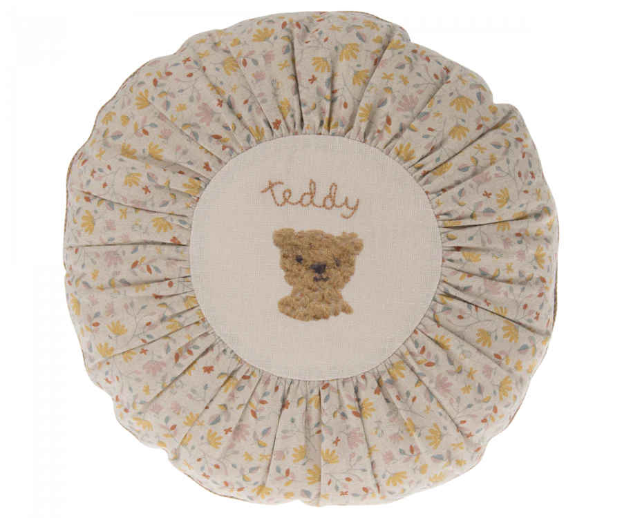 A round, floral-patterned Maileg Cushion, Small with a central embroidery of a teddy bear's face and the word "teddy" stitched above it in gentle script.
