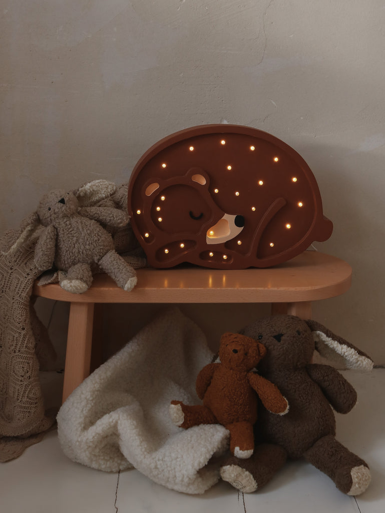 A cozy children's corner featuring a whimsical Handmade Bear Lamp with small glowing lights, surrounded by plush elephant toys and a soft blanket on a small wooden stool.