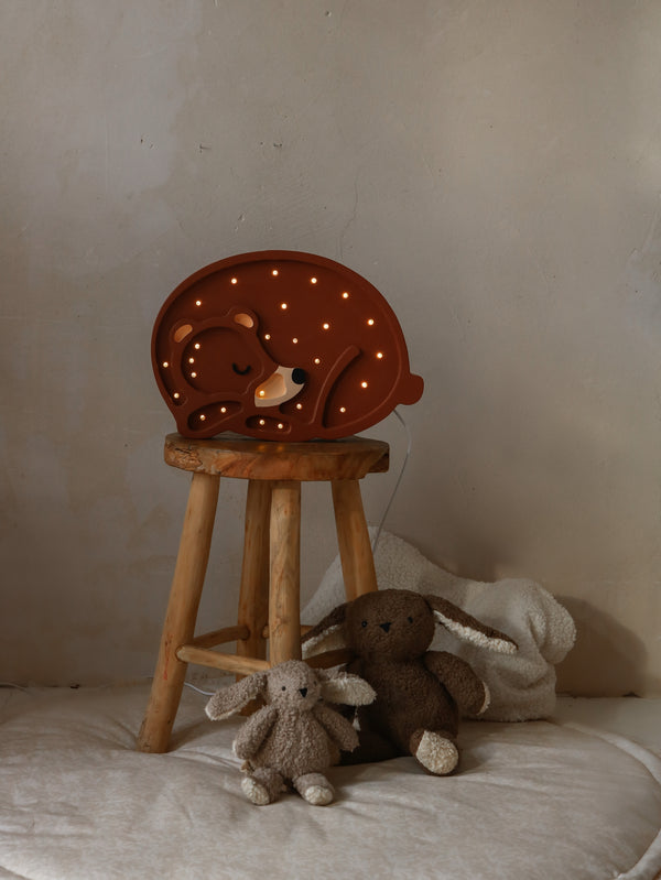 A cozy Handmade Bear Lamp in the shape of a sleeping bear sits atop a wooden stool, flanked by two plush teddy bears, against a muted backdrop.