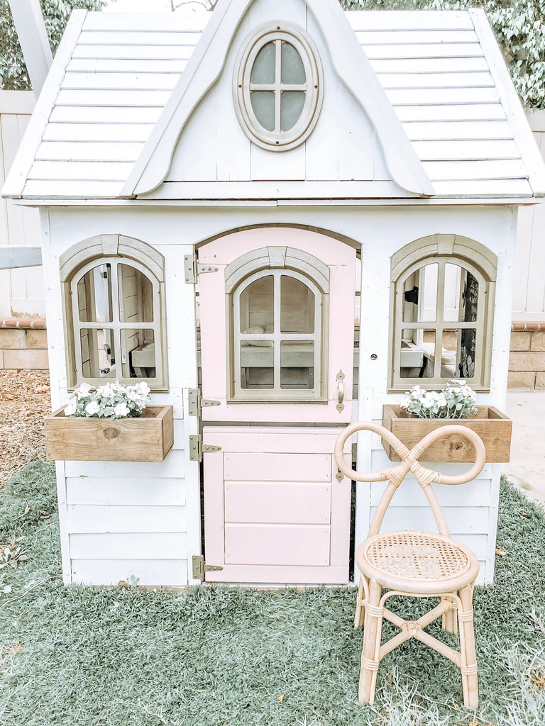 A quaint children's playhouse painted in white with pink doors and trim, featuring a round window on the upper facade and flower boxes under side windows, with a Toddler Rattan Bow Chair (Set of Two) in front.