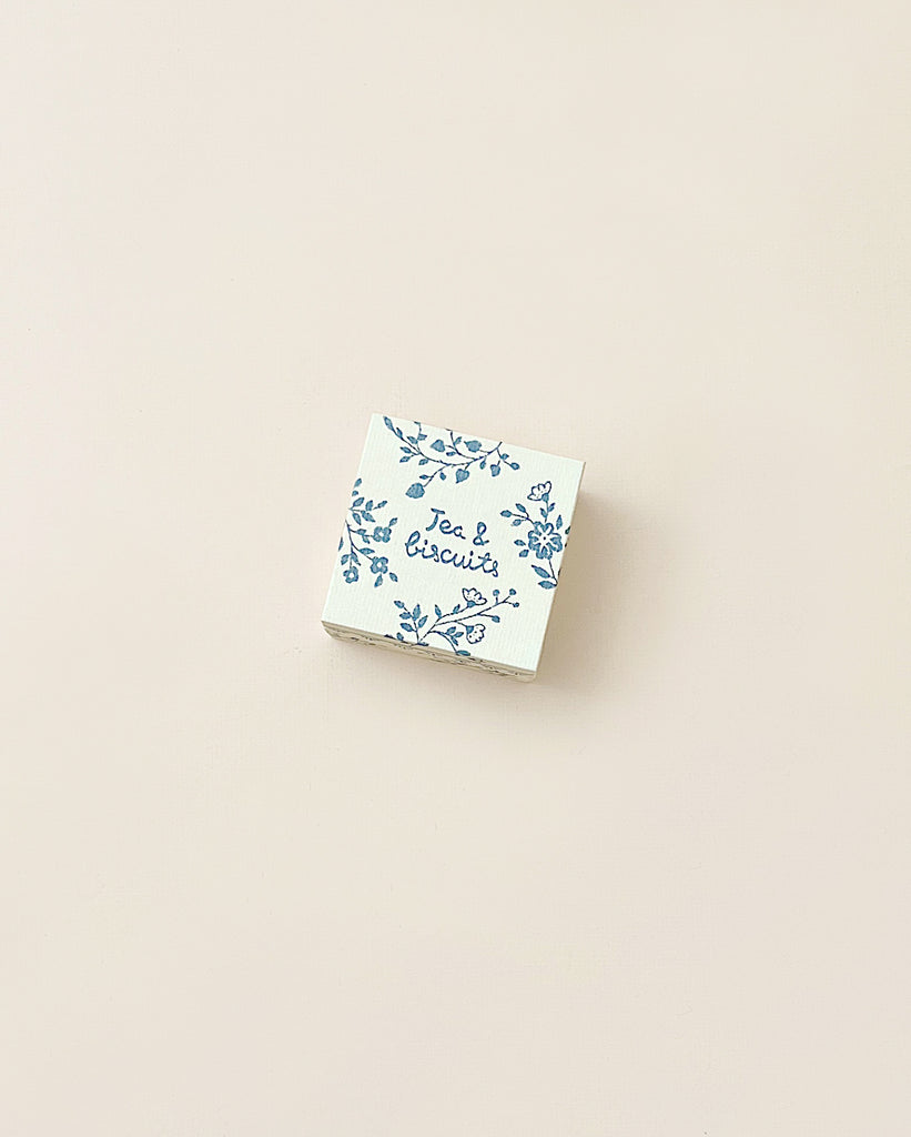 A small, square Maileg | Miniature Tea & Biscuits for Two box with a floral design and the words "tea & biscuits" printed on it, set against a plain, light beige background.