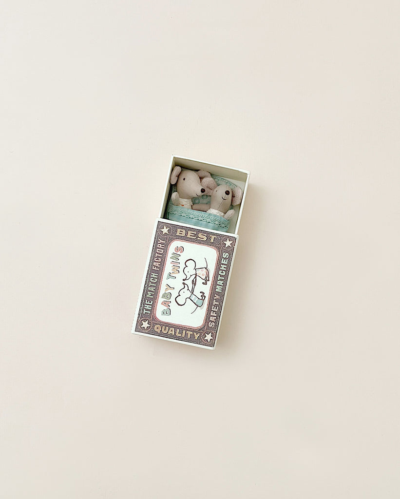A small, decorative Maileg | Twin Baby Mice In Matchbox with a "best quality" label containing two miniature plush bunny figurines peeking out, each dressed in a onesie, set against a light beige background.