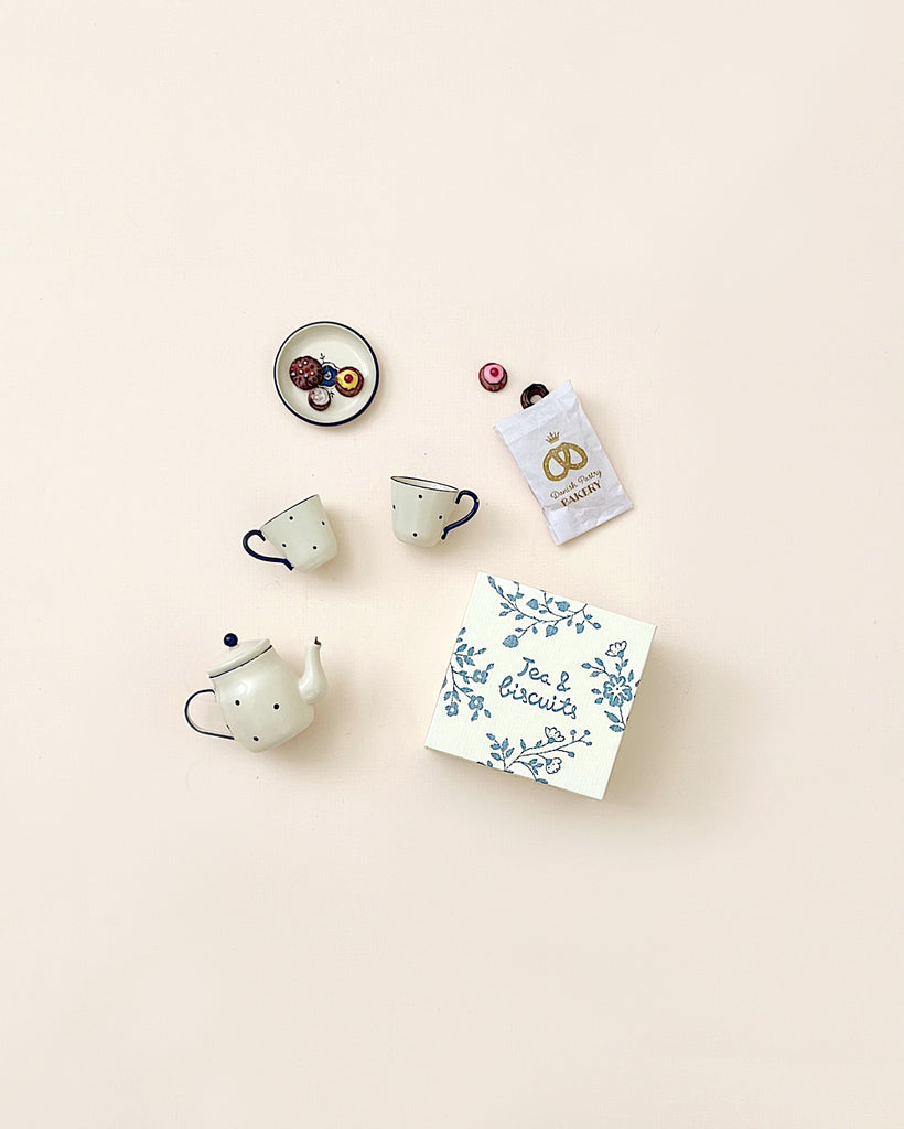 A neatly arranged Maileg Miniature Tea & Biscuits for Two set on a beige background, including a teapot, two cups, a plate with tiny cookies, and a packet labeled "tea biscuits.