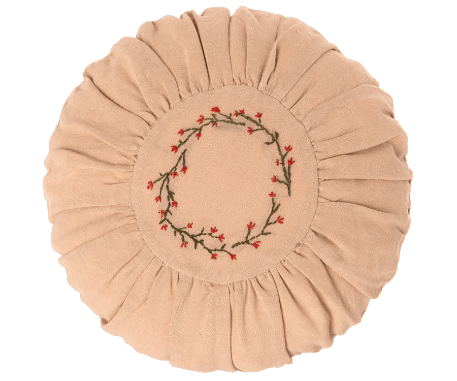 A round, beige Maileg Cushion, Small with a pleated edge and a center embroidered with a red and green floral wreath design, set against a black background.