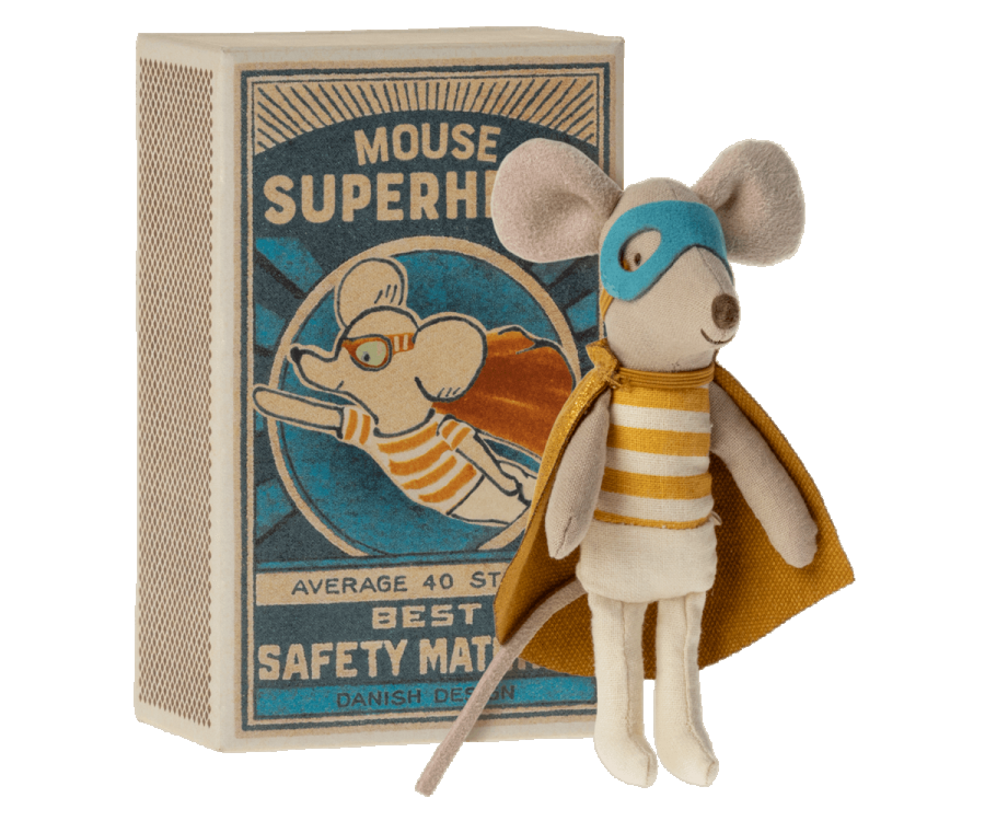 A Maileg | Superhero Mouse, Little Brother plush toy mouse dressed as a superhero with a cape and eye mask, standing beside a vintage book titled "Mouse Superhero." The toy and book display a charming, whimsical