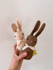 A hand holds two Polka Dot Club Rabbit Rattle toys, one beige and one brown, both adorned with organic cream neckties around their necks, against a plain light background.