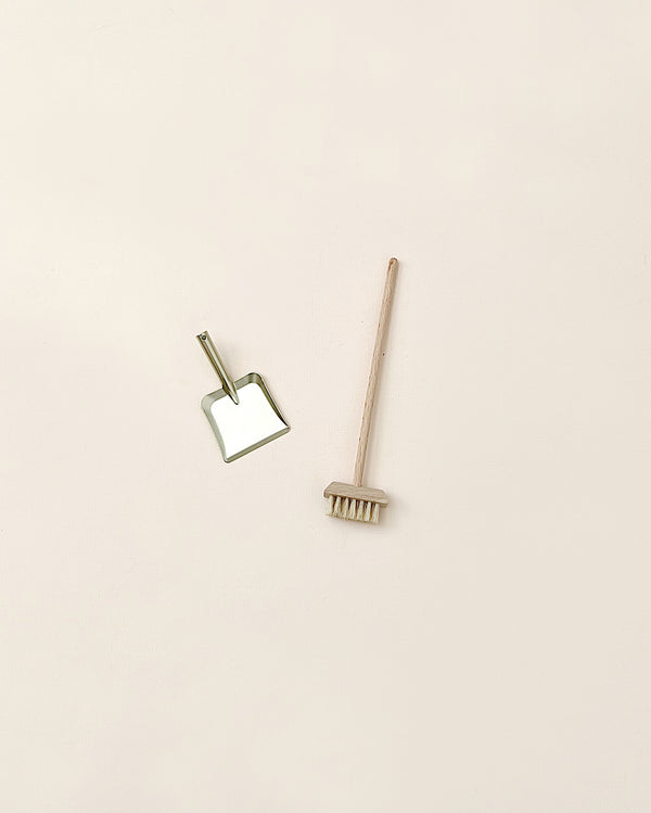 A minimalist image featuring a bamboo toothbrush lying horizontally and a small mirror with a gold frame set at an angle, alongside a Maileg Miniature Broom Set on a light pink background.