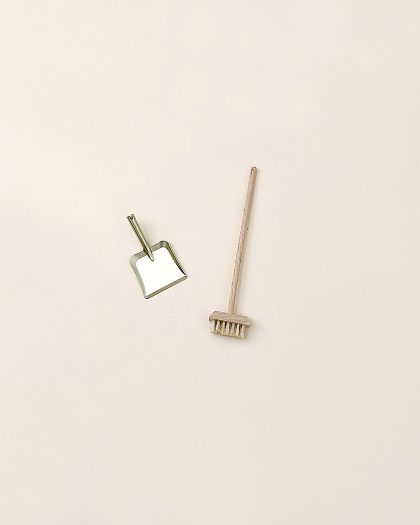 A minimalist image featuring a bamboo toothbrush lying horizontally and a small mirror with a gold frame set at an angle, alongside a Maileg Miniature Broom Set on a light pink background.
