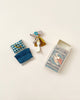 A small Maileg | Superhero Mouse, Little Brother figurine, wearing a cape and mask, stands next to folded textiles and a colorful matchbox with "superhero safety matches" written on it, arranged on a light.