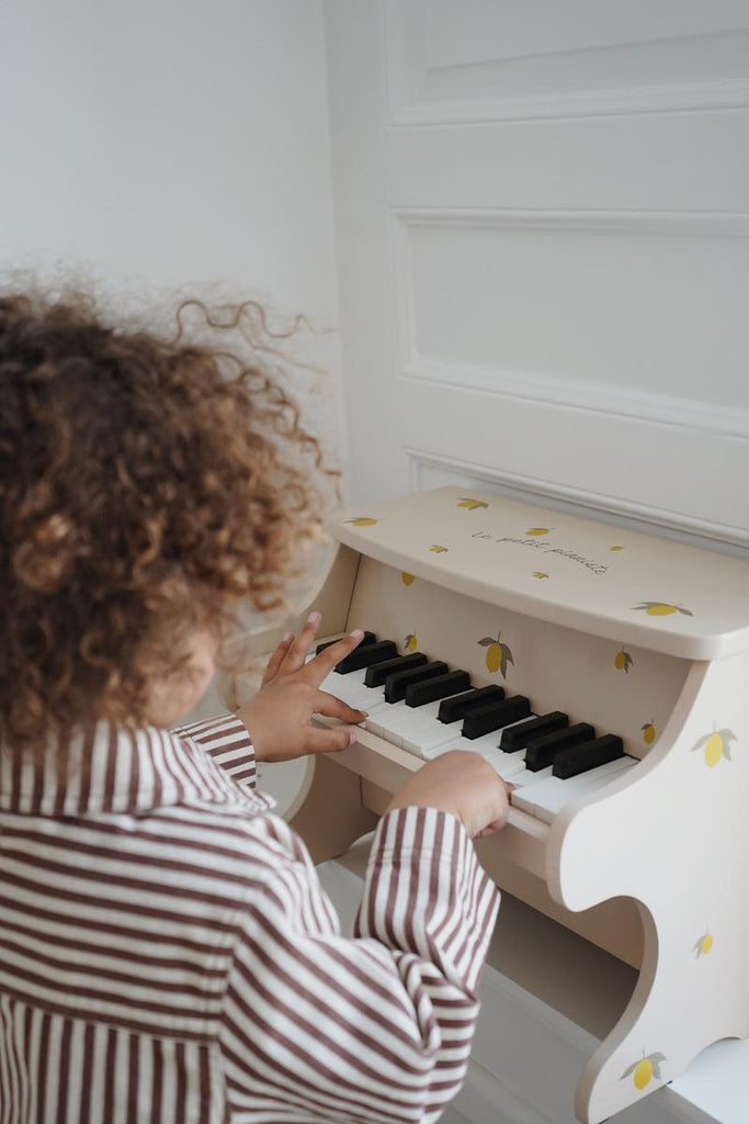A curly-haired child, donning a striped outfit, is playing a Wooden Toy Piano - Final Sale adorned with yellow lemon illustrations. The little pianist is facing away from the camera, and the white background contributes to a bright and minimalist setting.