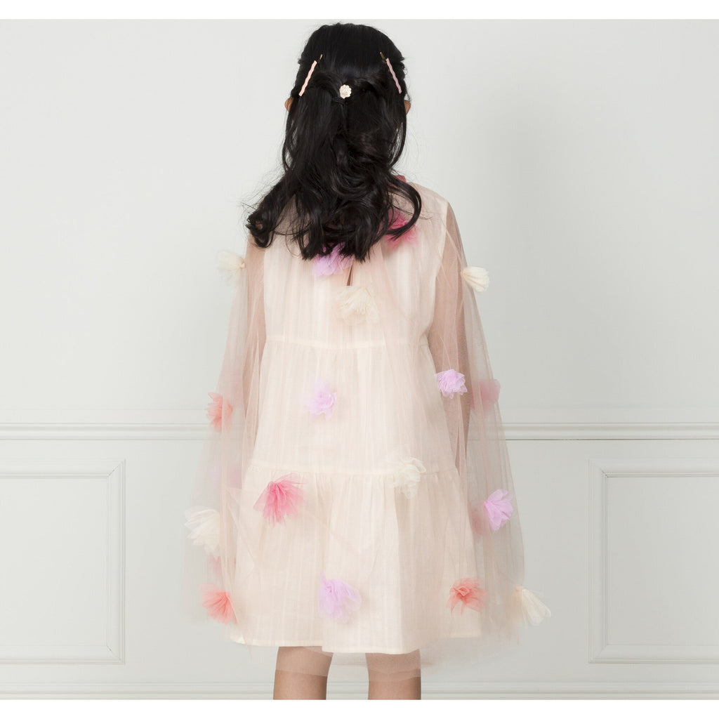 A young girl with her back to the camera wears a Meri Meri Flower Cape adorned with colorful tulle flowers, standing against a plain white wall.