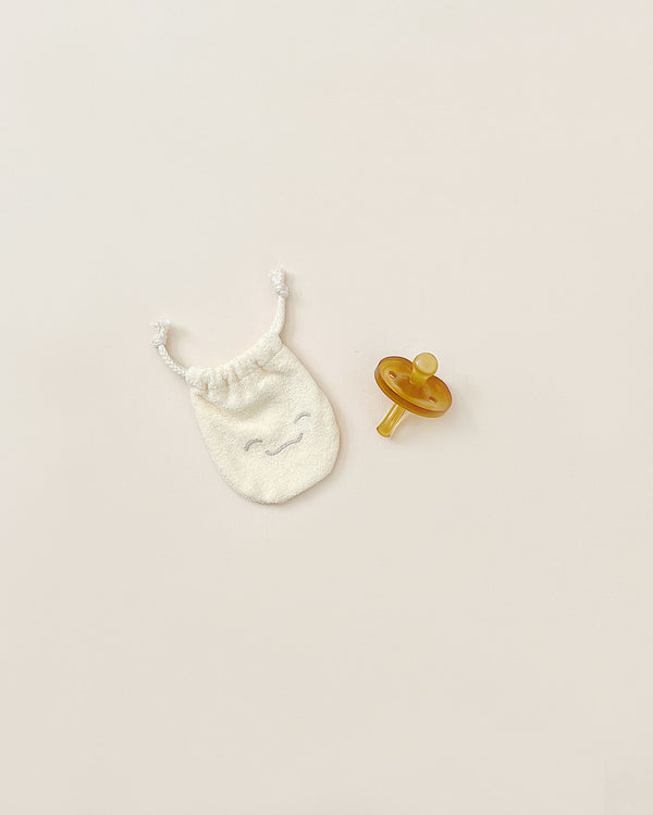 A small, smiley-faced white drawstring bag alongside a yellow Natursutten Butterfly Pacifier | 0-6 Months, small, both placed on a pale background.