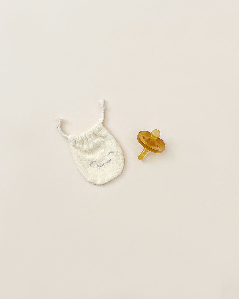 A small, smiley-faced white drawstring bag alongside a yellow Natursutten Butterfly Pacifier | 0-6 Months, small, both placed on a pale background.