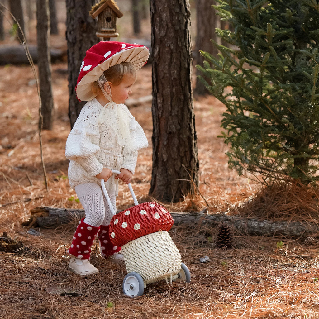 A toddler in a white sweater and red polka-dot pants pushes an Olli Ella Rattan Mushroom Luggy - Red toy with wheels through a pine forest, wearing a matching red and white polka-dot hat.