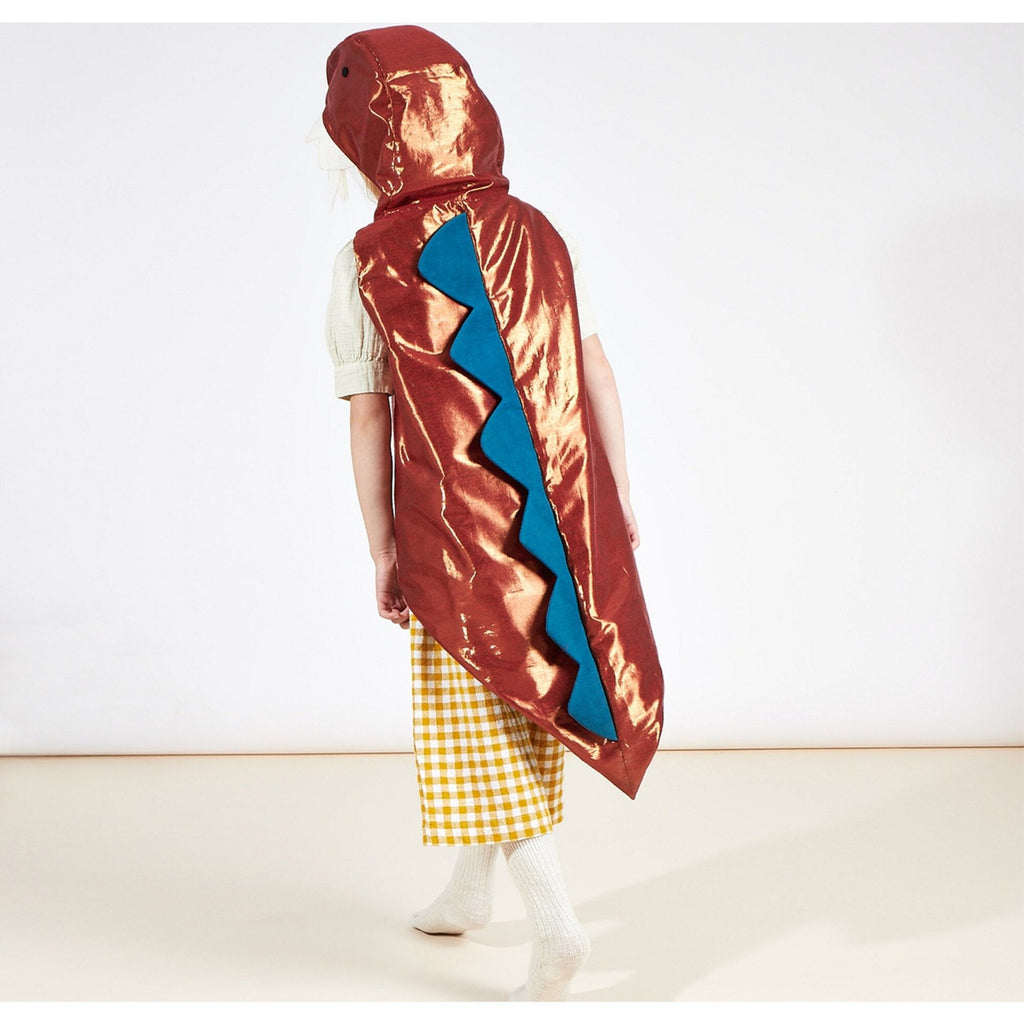 A child wearing a shiny red Meri Meri Dinosaur Costume with a large blue zigzag design on the back, standing against a light background. The costume includes a hood resembling a dinosaur head.