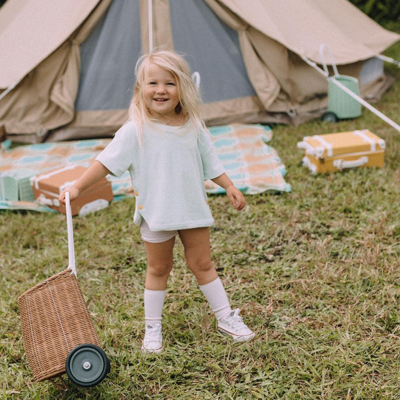 A young girl with blonde hair happily standing in a grassy field, holding an Olli Ella Rattan Luggy Basket, with a canvas tent in the background. She wears a white t-shirt.