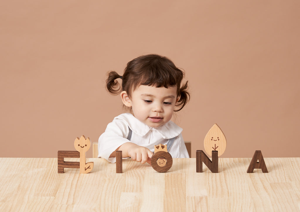 A toddler with pigtails plays with the Ultimate Wooden Alphabet Puzzle spelling "fiona" on a table, with cute animal designs on some letters, against a soft beige background.
