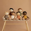 Six diverse posable Olli Ella Dinkum Dolls with different skin tones and hairstyles sitting on a small white bench against a beige background.