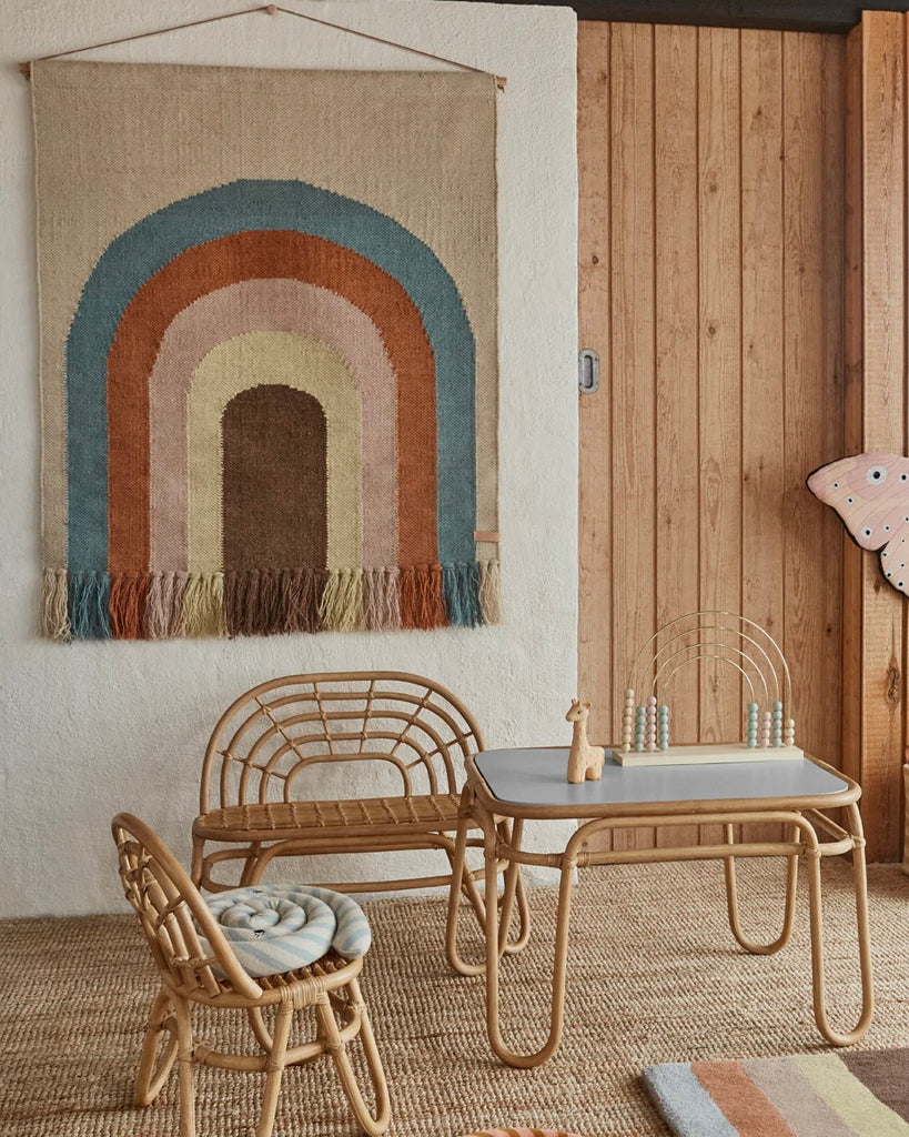 A cozy corner featuring a Follow The Rainbow Wall Rug on the floor, with a wooden table and rattan chairs on a textured rug. Soft natural lighting enhances the warm, inviting atmosphere.