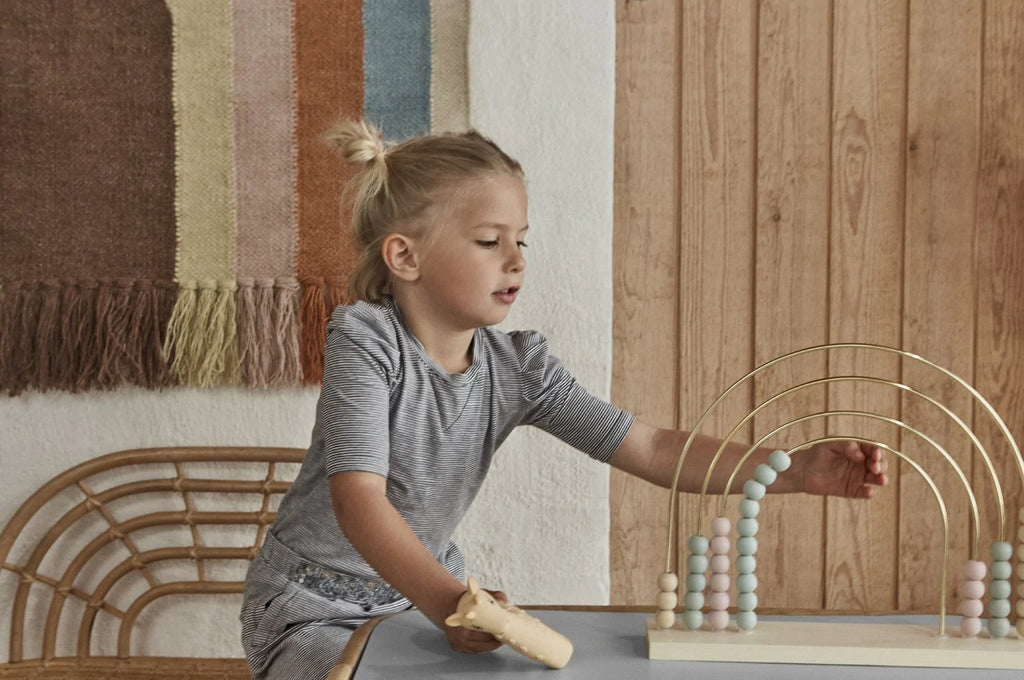 A young girl plays with a Follow The Rainbow Wall Rug at a table, in a room with wood paneling. She's focused on placing a piece on a circular toy structure.