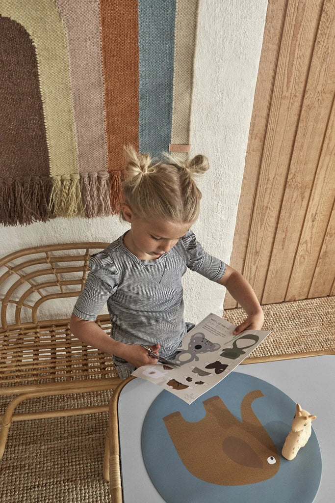A young girl with her hair in twin buns sits at a small table, reading a children's book. A Follow The Rainbow Wall Rug hangs in the background as wall decoration, and a figurine of a cat