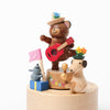 A whimsical display of the Mini Wooden Party Music Box featuring a smiling bear playing a red guitar, wearing a colorful hat, accompanied by a small dog and a mouse, each adorned with playful accessories, against.