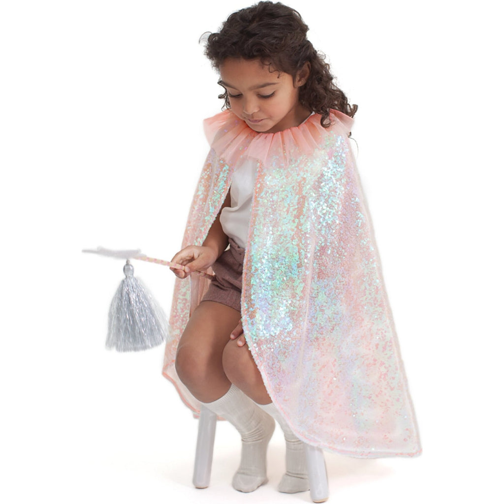 A young girl dressed as a fairy wearing a Meri Meri Iridescent Sequin Cape Costume and holding a sequin star wand, looks thoughtfully at glitter on her hand, against a white background.