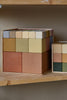 A collection of Raduga Grez Big Cube Block Sets in various muted colors, neatly stacked in a square formation, displayed on a wooden shelf.