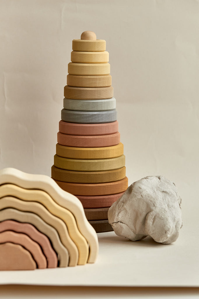 A minimalist display featuring a Raduga Grez | Handmade Large Pyramid Tower Stacker - Natural handcrafted by Raduga Grez in various earth tones beside a smaller, similarly shaped object and a white, textured stone on a neutral background.