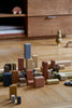 A Raduga Grez Extra Large Building Blocks Set is displayed on a wooden floor. This hand-crafted block set features buildings of various heights and shapes, all painted with non-toxic water-based paint. Nearby, a small wooden tree and minimalistic wooden vehicles invite open-ended play.