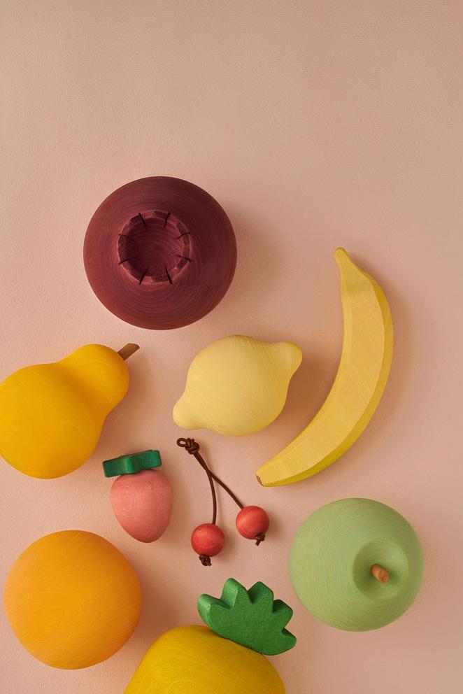 A collection of Raduga Grez Handmade Painted Wooden Fruits arranged on a light pink background. There are bananas, apples, a strawberry, cherries, a pear, and a lemon, creating a playful and artistic display.