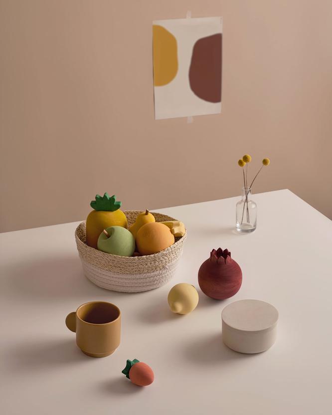 A still life composition on a neutral-toned table featuring a wicker basket filled with Raduga Grez Handmade Painted Wooden Fruits, two ceramic cups, a box, a pomegranate, and a small decor piece.