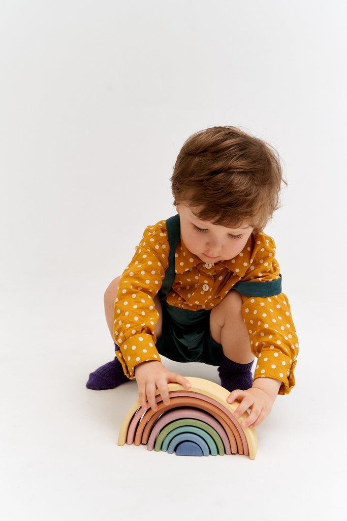 A young child plays with a colorful Raduga Grez 9-Piece Rainbow Stacker made of linden wood on a white background, wearing a polka dot shirt and overalls.