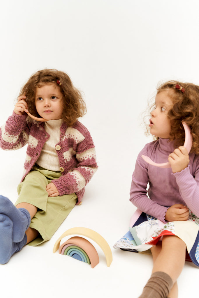 Two young girls with curly hair sit on the floor, one in a patterned sweater and the other in a purple top, both looking surprised while playing with a Raduga Grez 9-Piece Rainbow Stacker.