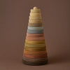 A Raduga Grez | Handmade Large Pyramid Tower Stacker - Natural composed of various-sized circular rings in gradient colors ranging from dark brown to light beige, neatly arranged to form a cone-like shape, coated with non-toxic paint, set against a