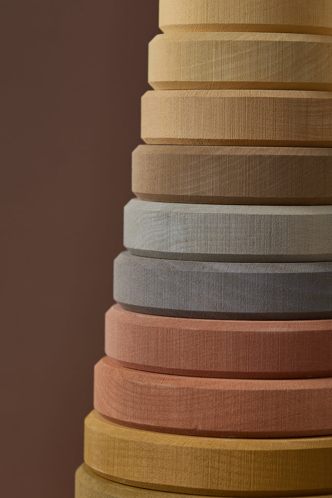 A stack of colorful fabric rolls in various shades including yellow, gray, and pink, arranged vertically against a soft brown background. The focus is on texture and color gradation, ideal for creating Raduga Grez | Handmade Large Pyramid Tower Stacker - Natural.