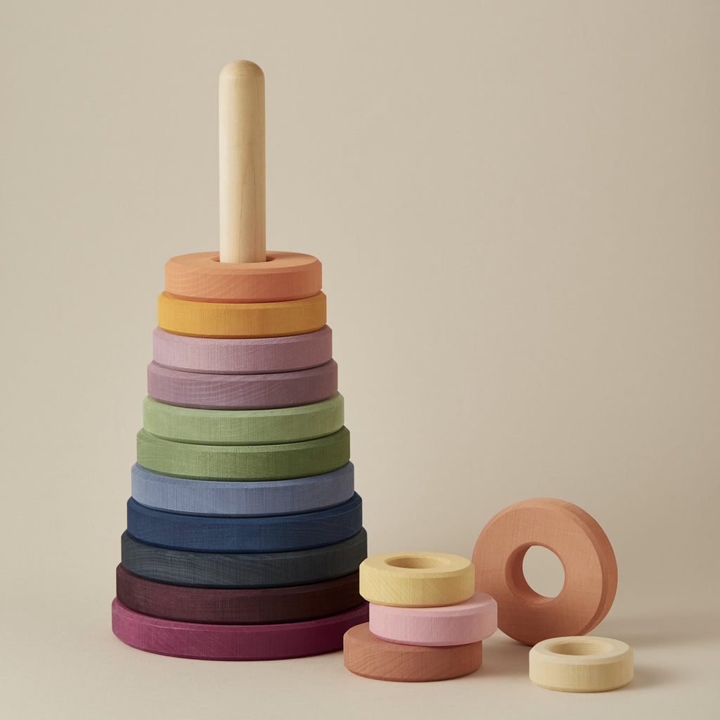 A Raduga Grez Large Pyramid Tower Stacker toy with a dowel, featuring rings in a gradient of colors from dark purple at the base to orange at the top, coated in non-toxic paint, set against a neutral