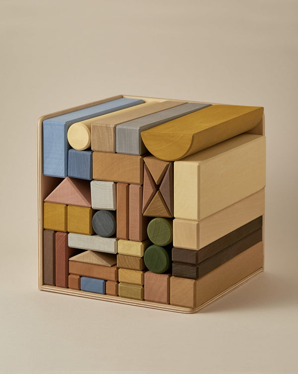 A neatly arranged stack of wooden blocks, various shapes, and colors, fitted into a square formation for creative play. The Raduga Grez | Big City Block Set includes rectangles, cylinders, and triangles in muted tones of yellow, blue, green, brown, and natural wood. The background is a plain beige.