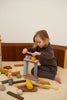 A young child with light brown hair sits on the floor in a cozy room, intently stacking colorful wooden blocks and balls from their Raduga Grez | Big City Block Set to create a structure. Toys are scattered around the child, and a paper-like accordion decoration is in the background, emphasizing creative play.