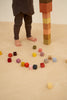 A child in brown trousers stands barefoot next to a tall stack of Raduga Grez Big Cube Block Set, with more vibrant cubes scattered around on a beige floor.