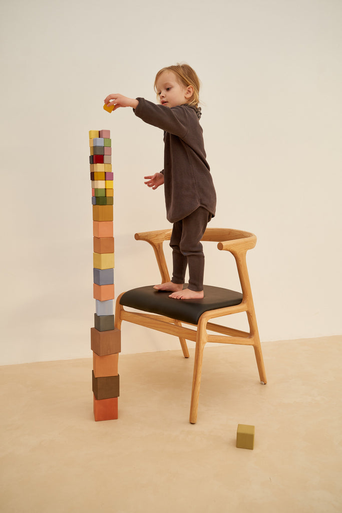 A toddler stands barefoot on a wooden chair, carefully placing a block from the Raduga Grez Big Cube Block Set on top of a tall tower of colorful cubes, with a plain backdrop.