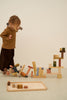 A young child with light brown hair, dressed in a brown top and dark brown pants, plays with an assortment of colorful wooden blocks, part of a Raduga Grez Extra Large Building Blocks Set. The child appears to be in the middle of a construction using the non-toxic water based paint blocks, and some have fallen to the floor.