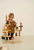 A young child with reddish-blonde hair, wearing a brown long-sleeved shirt, is focused on stacking a Raduga Grez Extra Large Building Blocks Set painted with non-toxic water-based paint into a tall, intricate tower. The background is a plain white wall and a light-colored floor. A tray with more blocks for open-ended play is partially visible.