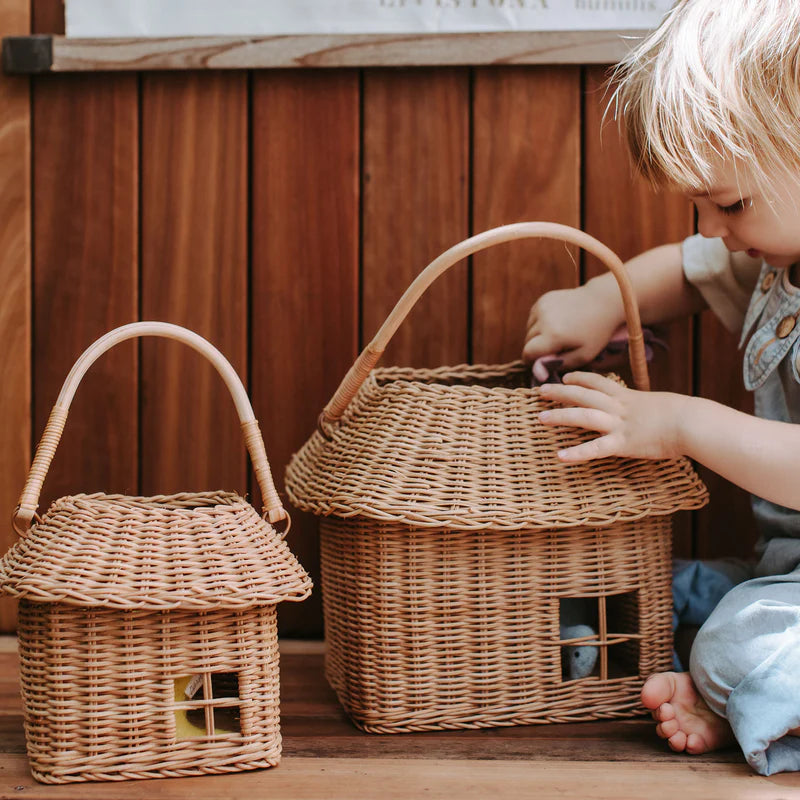 A young child sits beside two Olli Ella Rattan Hutch Baskets - Large that resemble houses, playing with a small object near one of them, set against a wooden background.