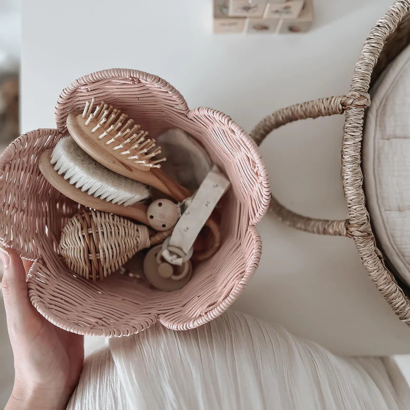 A person holding two Olli Ella Rattan Lily Baskets; the top basket is open, displaying various wooden hairbrushes and grooming tools inside. The setting has a soft, neutral color scheme.