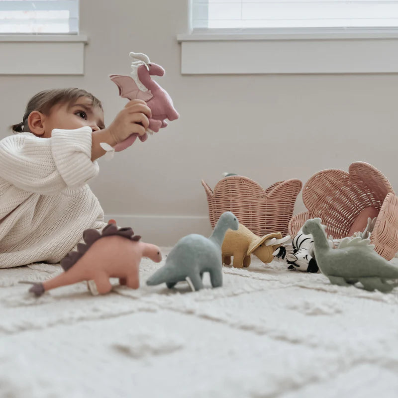 A young child plays on the floor with a variety of stuffed dinosaur toys, holding one up to examine it closely, surrounded by soft light filtering through the room and an Olli Ella Rattan Lily Basket Set nearby.